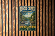A vintage-style metal sign for Rock Creek State Park in Iowa, featuring a scenic landscape with a waterfall and forest.