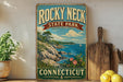 A vintage-style sign for Rocky Neck State Park in Connecticut, featuring a scenic landscape with a rocky coastline, a sailboat on the water, and a blue sky with clouds.