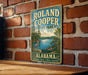 A vintage-style metal sign for Roland Cooper State Park in Alabama, featuring a scenic illustration of a lake, a crane, and a boat.