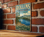 A vintage metal sign for Rollins State Park in New Hampshire, featuring a scenic image of a lake surrounded by mountains and pine trees.