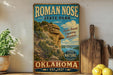 a vintage-style sign for Roman Nose State Park in Oklahoma, with a scenic mountain landscape and a blue sky in the background.