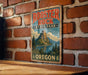 A vintage-style metal sign for Rooster Rock State Park in Oregon, featuring a mountain landscape and a lake, mounted on a brick wall.