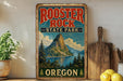 A vintage-style sign for Rooster Rock State Park in Oregon, featuring a mountain landscape and a lake, is displayed on a shelf next to a wooden cutting board and some lemons.