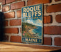 A vintage-style metal sign for Maine State Park, featuring a scenic coastal landscape with a rocky cliff, pine trees, and a beach.