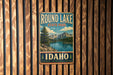 A vintage metal sign for Round Lake State Park in Idaho, featuring a scenic landscape with mountains, a lake, and pine trees.