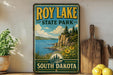 A vintage-style sign for Roy Lake State Park in South Dakota, featuring a scenic landscape with a lake, mountains, and pine trees.