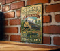 A vintage-style metal sign for Ryserson Station State Park in Pennsylvania, featuring an illustration of a historic building and a bridge.