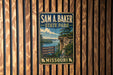A vintage metal sign for Sam A. Baker State Park in Missouri, featuring a scenic landscape with a cliff, water, and trees.
