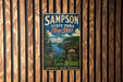 A vintage metal sign for Sampson State Park in New York, featuring a scenic landscape with a lake and mountains.