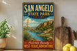 A vintage-style metal sign for San Angelo State Park in Texas, featuring a scenic landscape with a river, mountains, and desert vegetation.