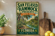 A vintage-style sign for San Francisco Hammock Preserve, featuring a tropical landscape with palm trees and a river, is displayed on a shelf next to a wooden cutting board and some lemons.