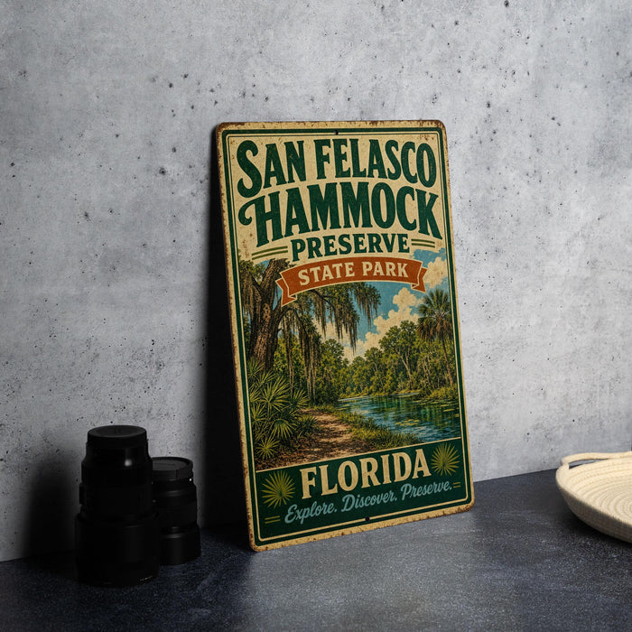 A vintage-style metal sign for San Felasco Hammock Preserve, featuring a tropical landscape with palm trees and a river, is displayed on a gray concrete surface.