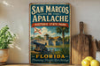 A vintage-style sign for San Marcos, Florida, featuring a historical state park and a cannon, with a wooden paddle and a potted plant in the background.