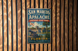 A vintage-style metal sign for San Marcos de Apalache Historic State Park in Florida, featuring a palm tree and a cannon, is displayed on a wooden wall.