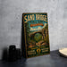A vintage-style metal sign for Sand Bridge State Park in Pennsylvania, featuring a scenic illustration of the bridge surrounded by lush greenery.
