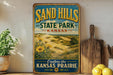 A vintage-style sign for Sand Hills State Park in Kansas, featuring a scenic landscape with dunes and flowers, is displayed on a shelf next to a wooden cutting board and some lemons.