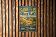A vintage-style metal sign for Sand Hills State Park in Kansas, featuring a scenic landscape with sunflowers and a message encouraging visitors to explore the Kansas prairie.