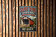 A vintage-style metal sign for the Sandy Creek Covered Bridge in Missouri, featuring an illustration of a covered bridge and the text "Sandy Creek Covered Bridge Missouri".