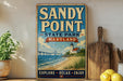 A vintage-style sign for Sandy Point State Park in Maryland, featuring a lighthouse and a beach scene, is displayed on a shelf next to a wooden cutting board and some lemons.