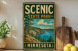 A vintage-style sign for Scenic State Park in Minnesota, featuring a scenic landscape with a lake, mountains, and pine trees.