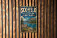 A vintage-style metal sign for Scofield State Park in Utah, featuring a scenic mountain landscape with a lake and a boat.