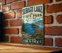 A vintage metal sign for Sebago Lake State Park in Maine, featuring a scenic landscape with a lake, mountains, and pine trees.