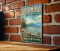A vintage-style metal sign for Sequim Bay State Park in Washington, featuring a scenic landscape with mountains, pine trees, and a lake.