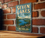 A vintage metal sign for Seven Lakes State Park in Michigan, featuring a scenic landscape with a lake surrounded by mountains and pine trees.