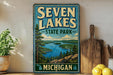 A vintage-style sign for Seven Lakes State Park in Michigan, featuring a scenic landscape with a lake surrounded by pine trees and mountains.