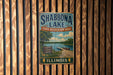 A vintage metal sign for Shabbona Lake, Illinois, featuring a scenic illustration of the lake and surrounding landscape.