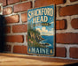 A vintage metal sign for Shackford Head State Park in Maine, featuring a scenic coastal landscape with rocky cliffs, trees, and a body of water.
