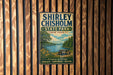 A vintage-style metal sign for Shirley Chisholm State Park in New York, featuring a scenic landscape with a lake, trees, and a picnic table.
