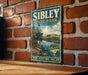 A vintage-style metal sign for Sibley State Park in Minnesota, featuring a scenic landscape with a lake and mountains.