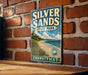A vintage-style metal sign for Silver Sands State Park in Connecticut, featuring a scenic beach landscape with a wooden fence and a bird in flight.