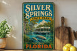 A vintage-style sign for Silver Springs State Park in Florida, featuring a scenic image of a boat on a river surrounded by palm trees and other natural elements.