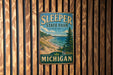 A vintage metal sign for Sleeper State Park in Michigan, featuring a scenic landscape with a beach, mountains, and pine trees.