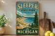 A vintage-style sign for Sleeper State Park in Michigan, featuring a scenic landscape with a beach, mountains, and pine trees.