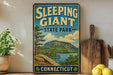 A vintage-style sign for Sleeping Giant State Park in Connecticut, featuring a scenic mountain landscape with a lake and pine trees.