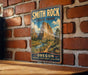 A vintage-style metal sign for Smith Rock State Park in Oregon, featuring a scenic landscape with mountains and a winding path.