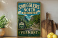 A vintage-style sign for Smugglers' Notch State Park in Vermont, featuring a scenic mountain landscape with a winding road.