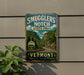 A vintage-style metal sign for Smugglers' Notch State Park in Vermont, featuring a scenic mountain landscape with a winding road.