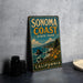 A vintage-style sign for Sonoma Coast State Park, featuring a scenic coastal landscape with mountains, ocean, and flowers.