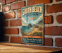 A vintage-style metal sign for South Beach State Park in Oregon, featuring a scenic coastal landscape with mountains, water, and beach.