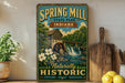 A vintage-style sign for Spring Mill State Park, Indiana, featuring a scenic illustration of a mill and waterfall.