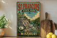 A vintage-style sign for St. Francis State Park in Missouri, featuring a scenic landscape with a river, trees, and mountains.