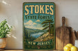 A vintage-style sign for Stokes State Forest in New Jersey, featuring a scenic landscape with pine trees, a lake, and mountains.