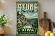 A vintage-style sign for Stone State Park, featuring a scenic landscape with a river and mountains, is displayed on a shelf next to a wooden cutting board and some potted plants.