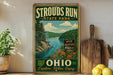 A vintage-style sign for Strouds Run State Park in Ohio, featuring a scenic landscape with a lake and mountains.