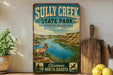 a vintage-style sign for Sully Creek State Park in North Dakota, with a scenic illustration of a river and mountains in the background.