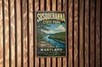 A vintage metal sign for Susquehanna State Park in Maryland, featuring a scenic landscape with a river and mountains.